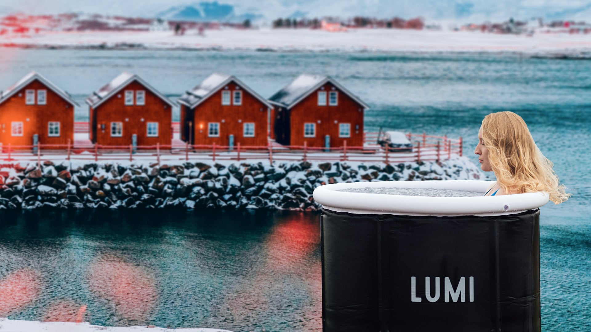 Image of a woman sat in a LUMI ice bath with a backdrop of the Baltic sea and snow covered red wooden houses