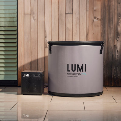 LUMI Cube Chiller positioned next to a large LUMI Recovery Pod against wooden wall paneling, showcasing compact portable cooling system for ice baths on light-colored tile flooring. Professional product photography demonstrates size comparison and sleek design.