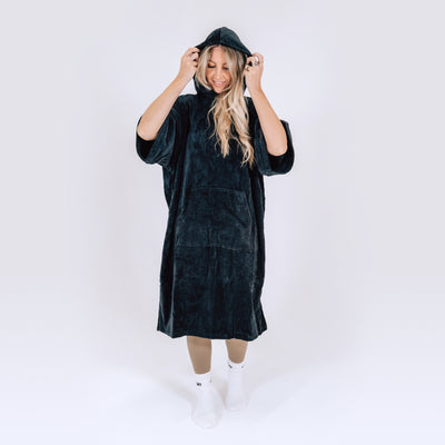 LUMI Towel Poncho shown in black plush cotton fabric, featuring oversized hooded design with front pocket, modeled against white background. Knee-length robe demonstrates roomy fit and cozy comfort perfect for post-swim or ice bath use.